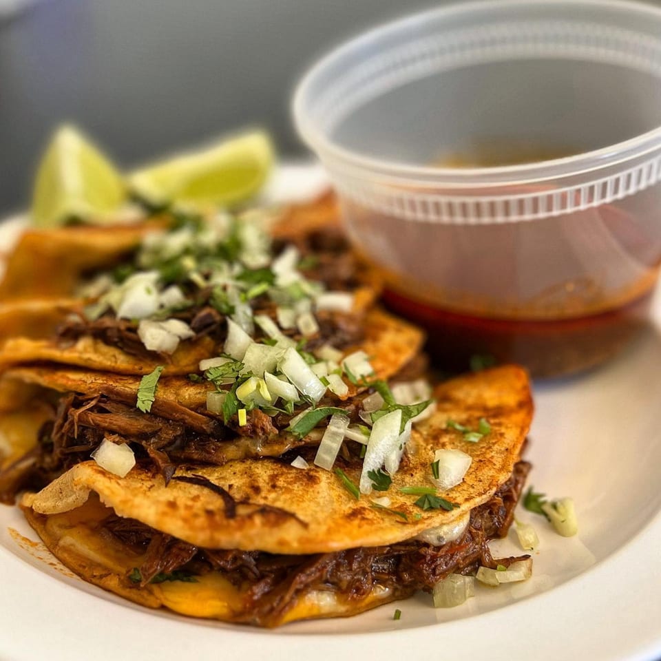 Three birria tacos with shredded meat, onions, cilantro, lime wedges, and a side of consommé.