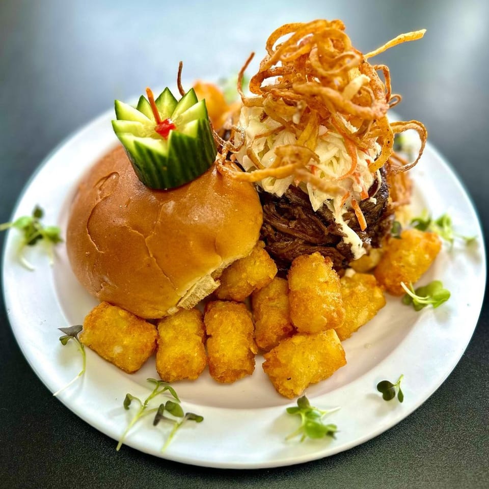 A Michigan Deli pulled pork sandwich with coleslaw, crispy onions, tater tots, and a cucumber garnish.