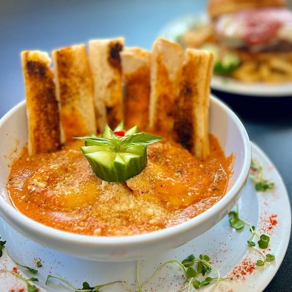 A bowl of ravioli in red sauce with parmesan cheese, garnished with cucumber and basil, served with breadsticks.